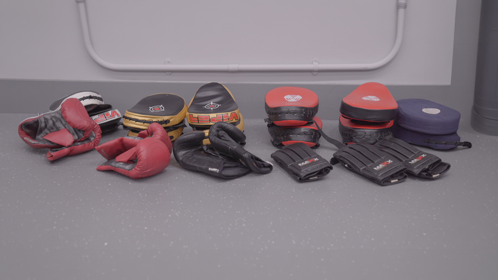 A collection of boxing gear, including red gloves, black and red focus mitts, and padded gloves, neatly arranged on a gray gym floor, suggesting readiness for training.