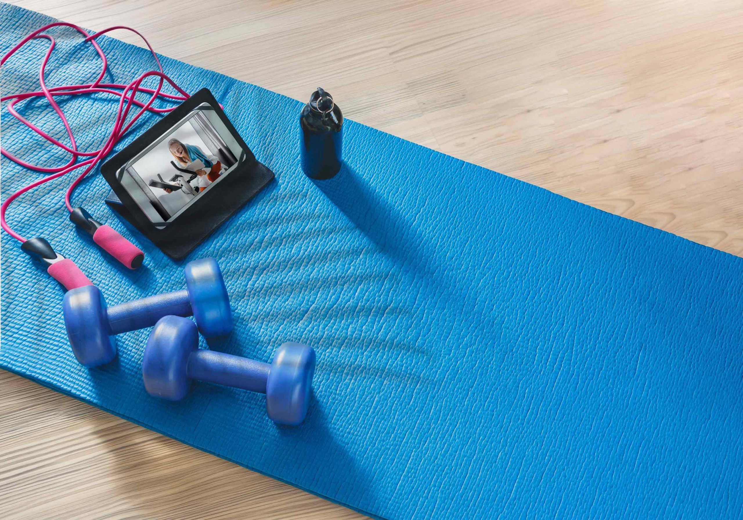 A blue exercise mat with two blue dumbbells, a pink jump rope, a tablet showing a fitness video, and a black water bottle on a wooden floor.