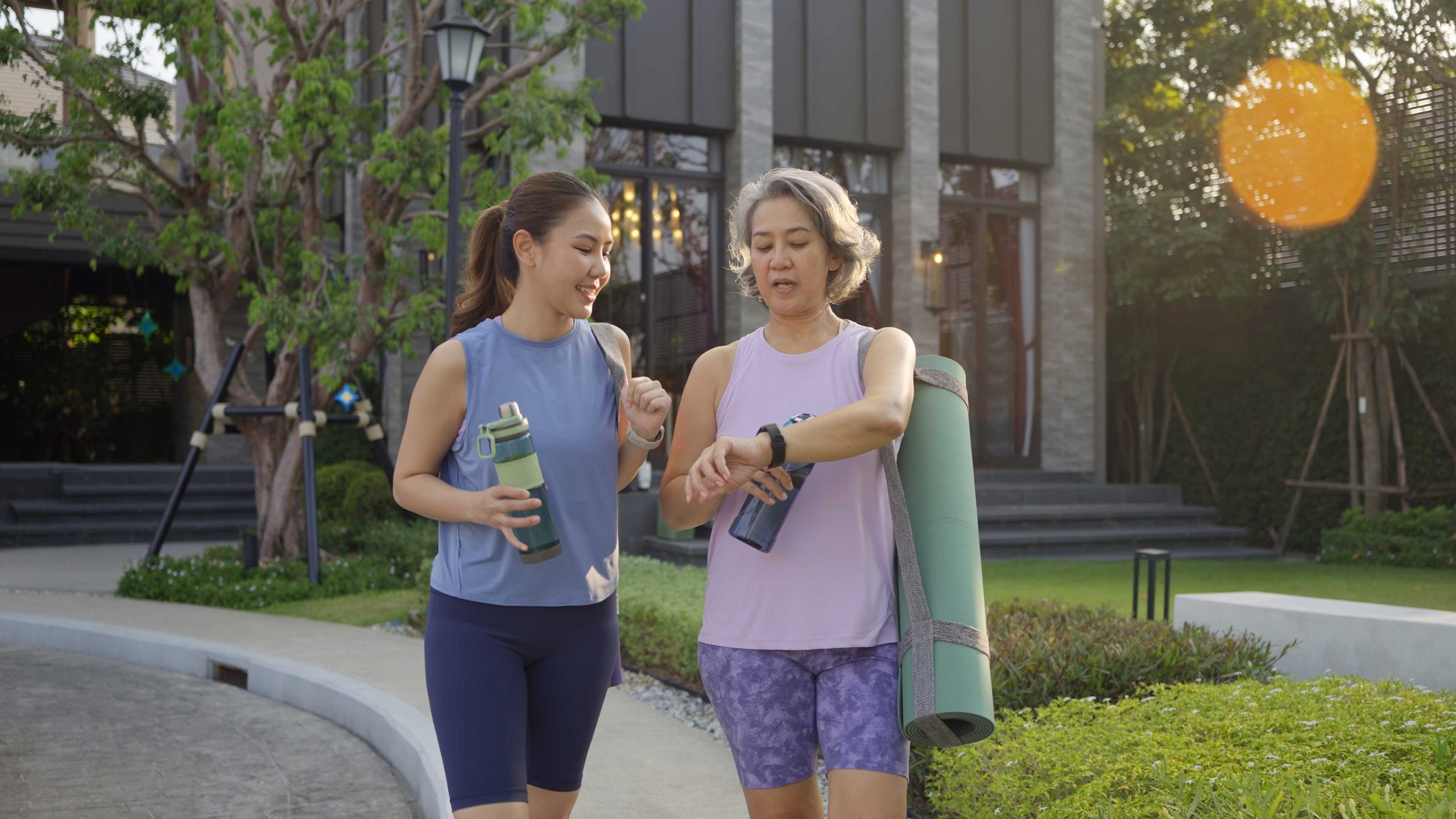 Two women in athletic wear walking outdoors with water bottles; one carries a yoga mat and checks her smartwatch.