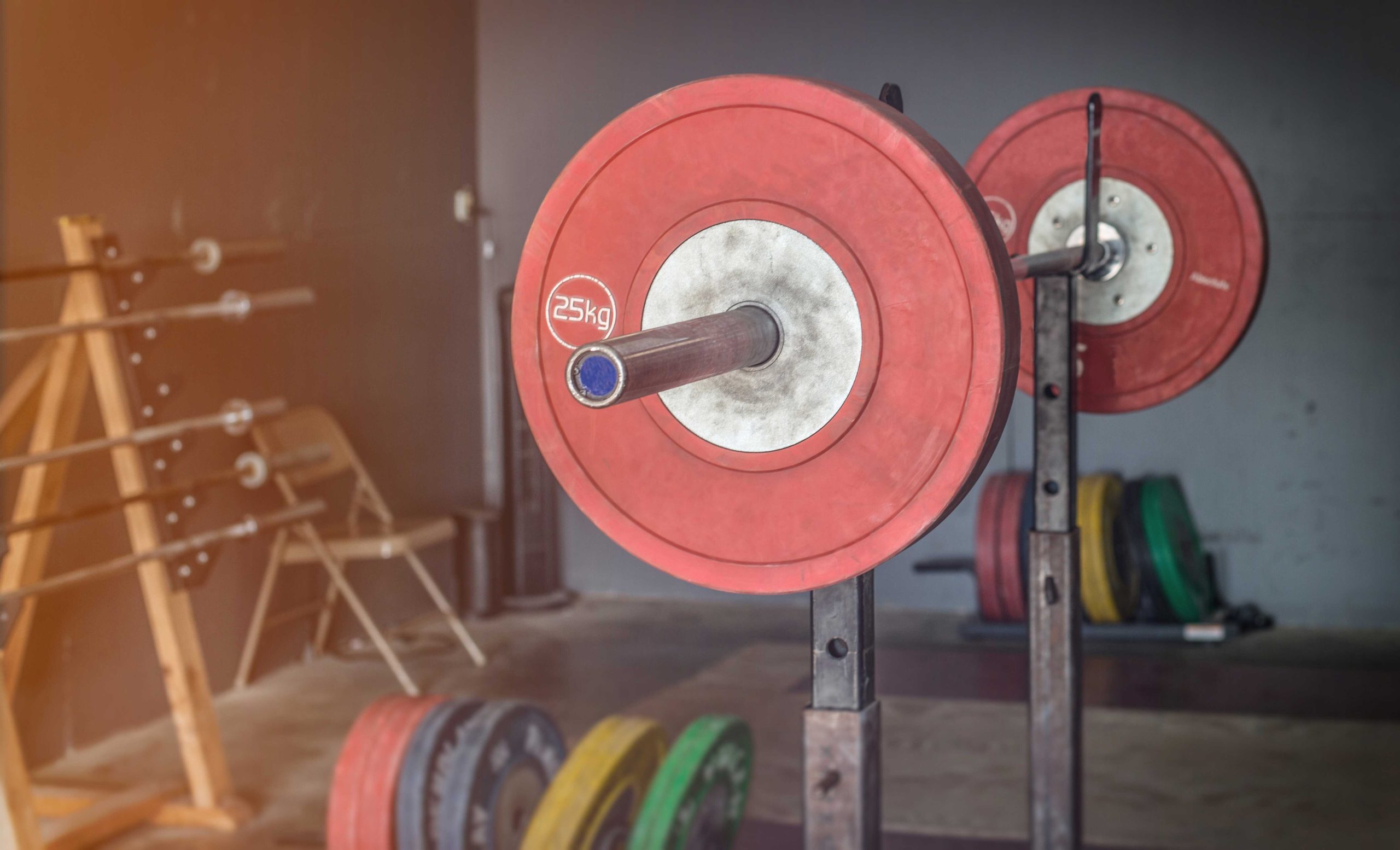 Barbell with 25kg red weight plates set on a squat rack in a gym with additional weight plates and chairs in the background.