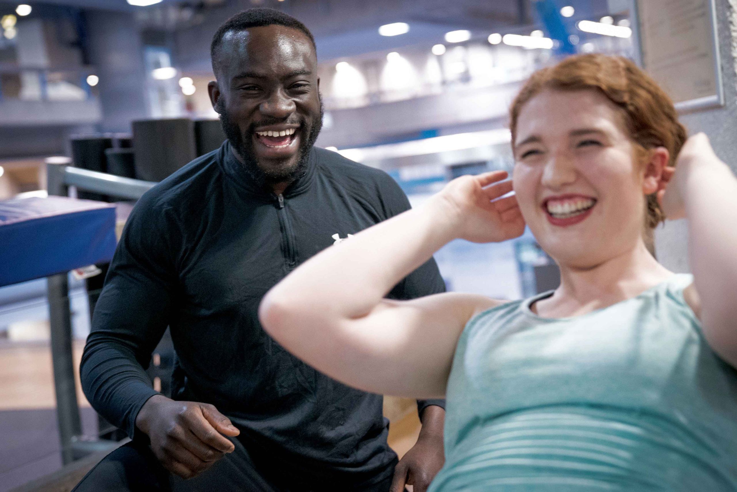 Trainer in black watches as a young man in a tank top performs sit-ups in a gym setting.