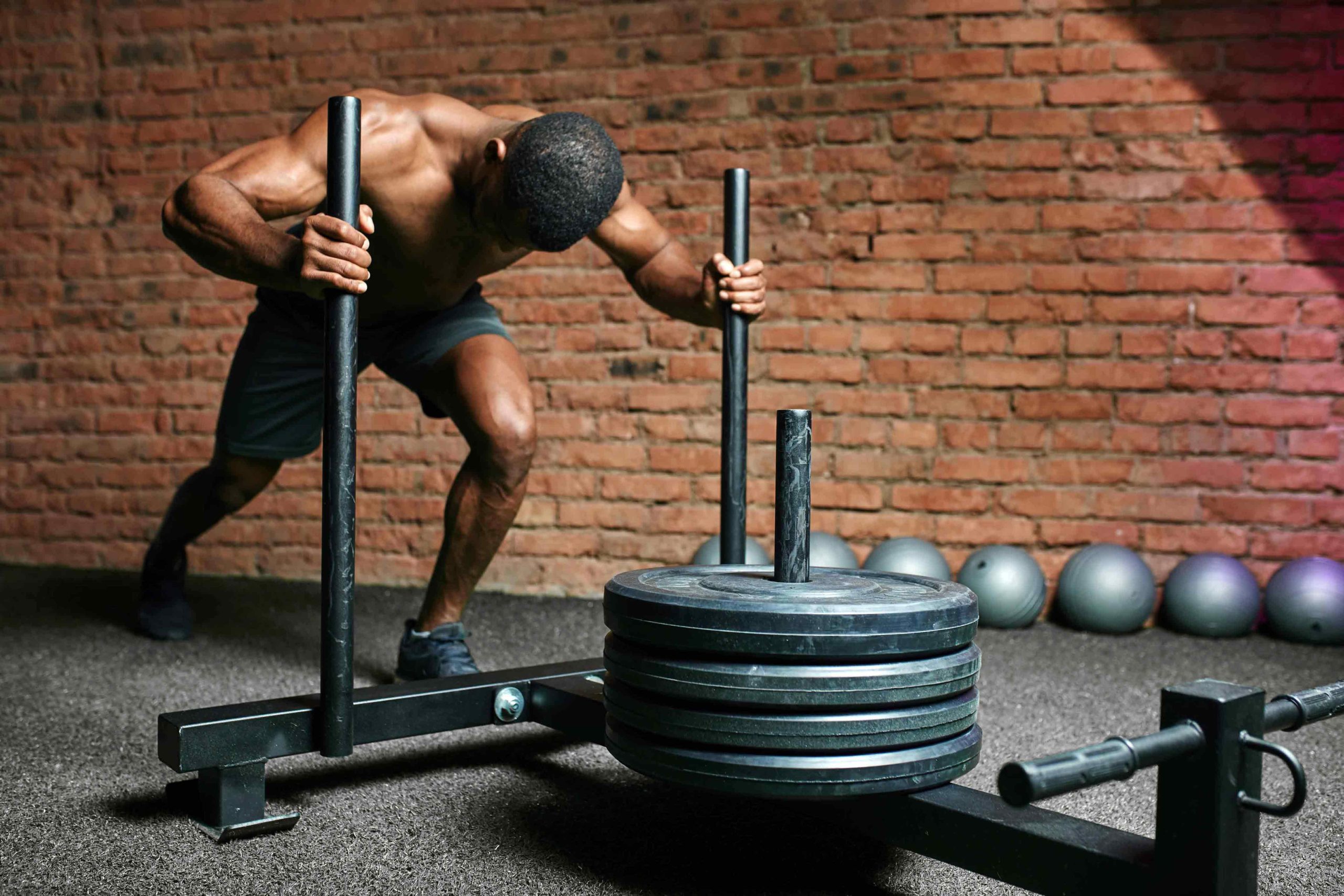 Athlete pushing a weighted sled on a textured gym floor, set against an exposed brick wall. The scene conveys strength and determination.