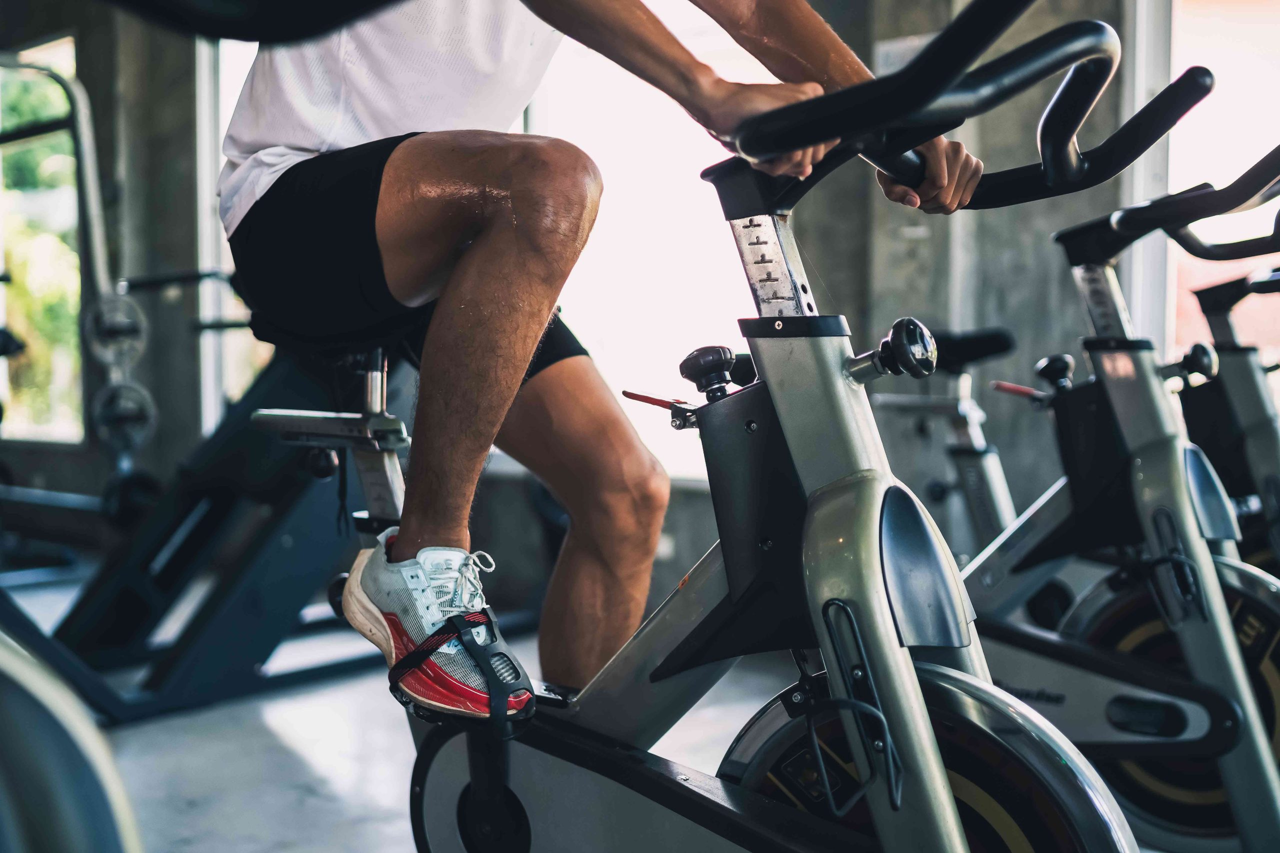 Person wearing white sneakers and black shorts exercising on a stationary bike in a gym with natural light.