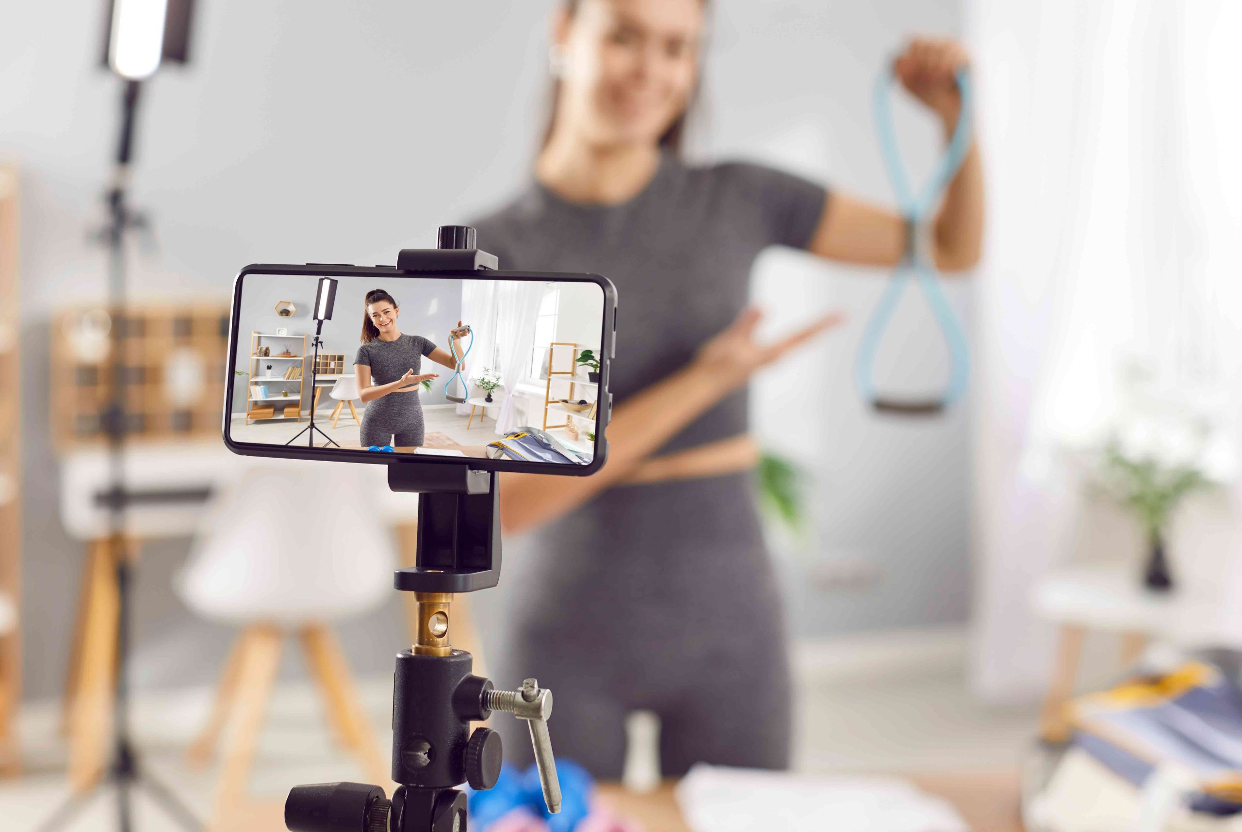 Person recording a fitness video at home, holding a blue resistance band in front of a smartphone mounted on a tripod.