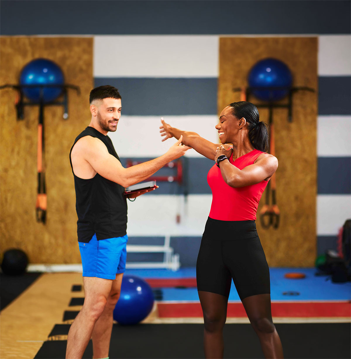 Trainer instructing a woman in a red top doing arm stretches in a gym environment with fitness equipment in the background.
