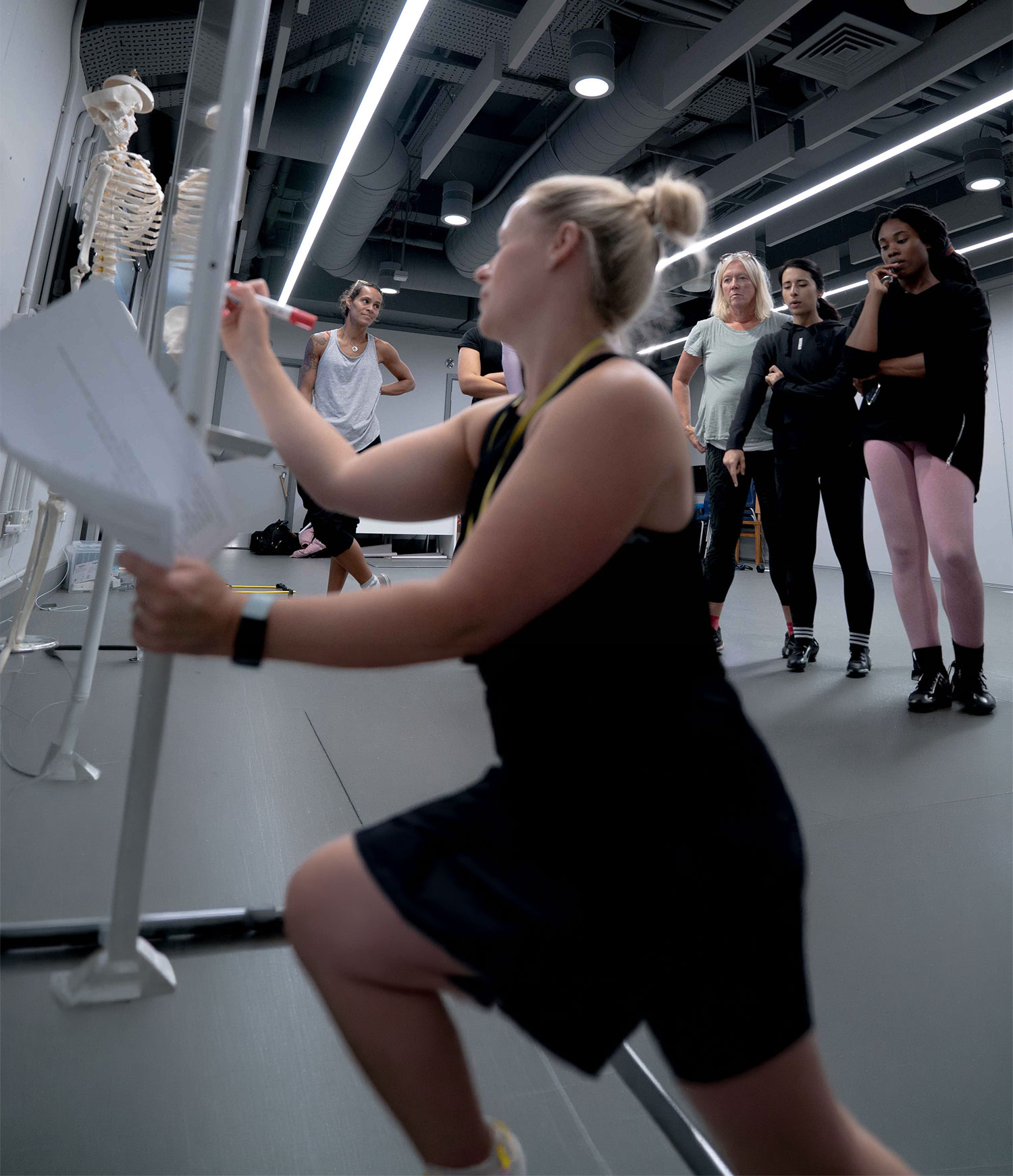 Group of people in a classroom observing as an instructor writes on a whiteboard beside a skeleton model.
