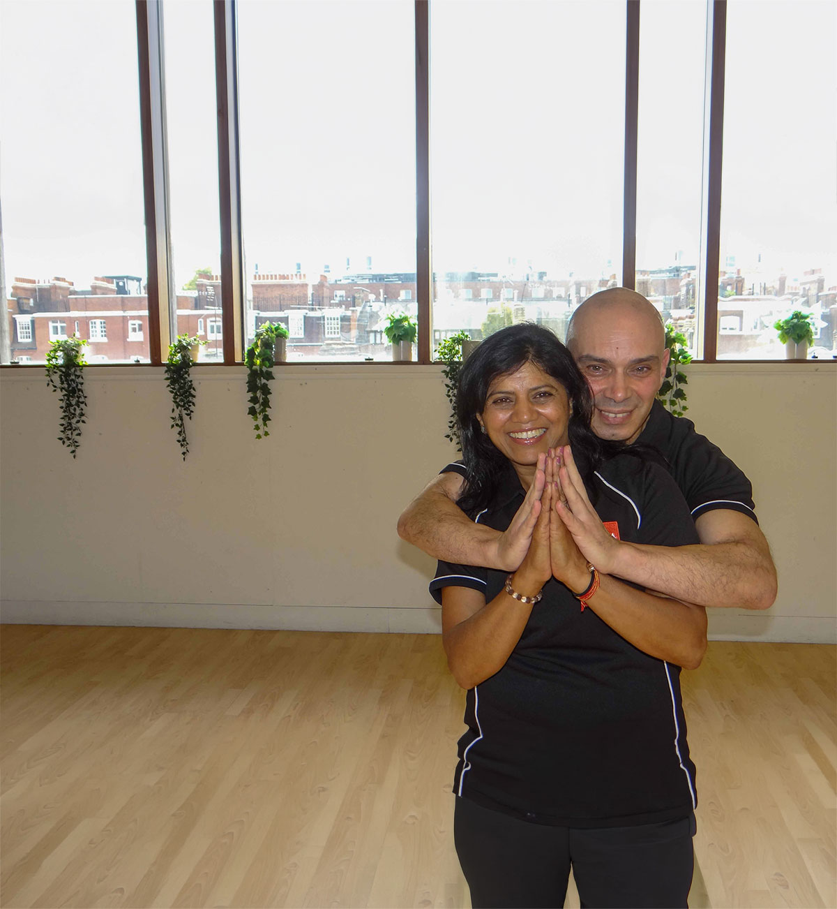 Two people in black shirts practicing a partner yoga pose indoors, with plants on a windowsill in the background.