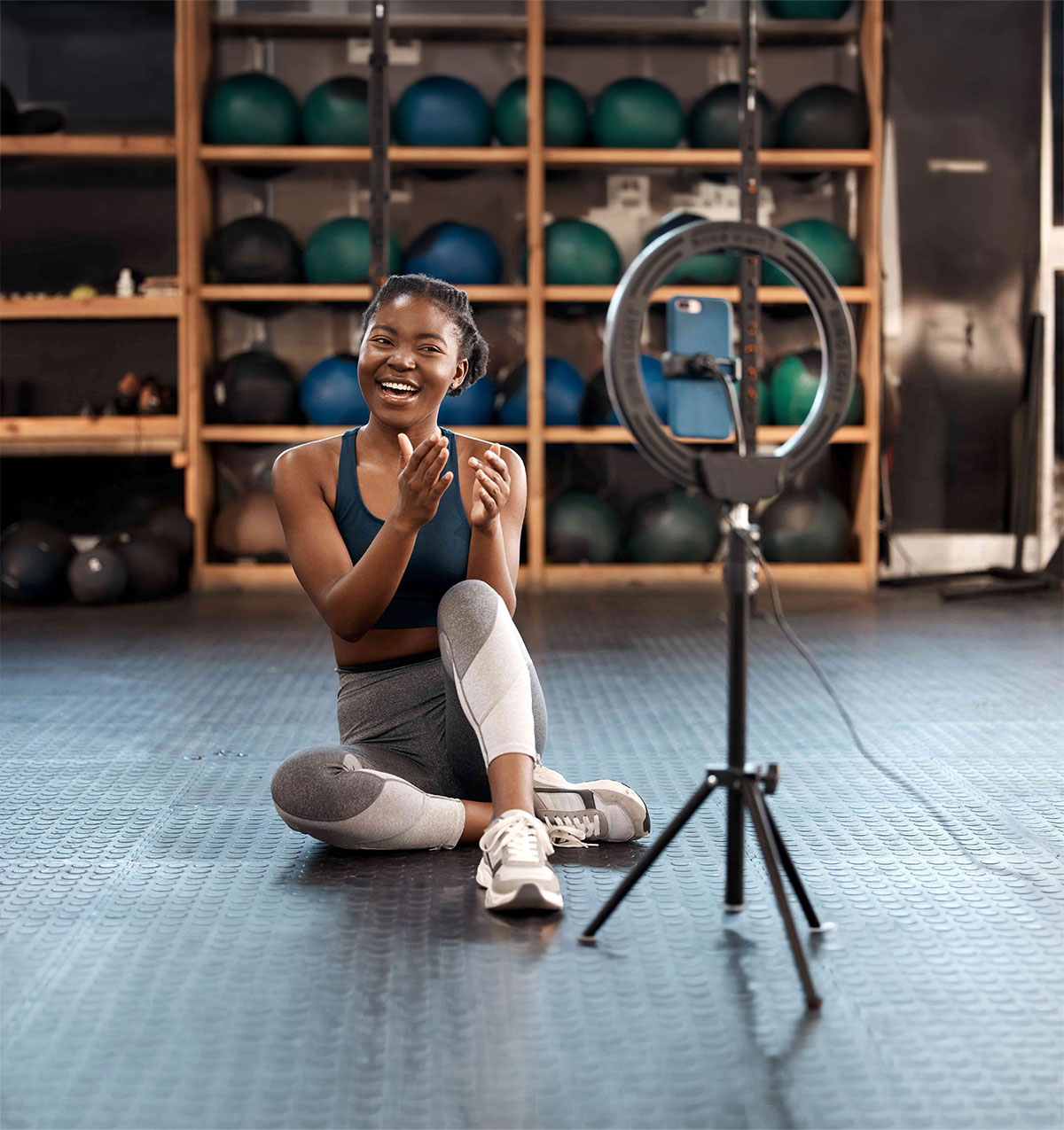 Woman in athletic wear sitting on gym floor and recording video with smartphone on ring light tripod.
