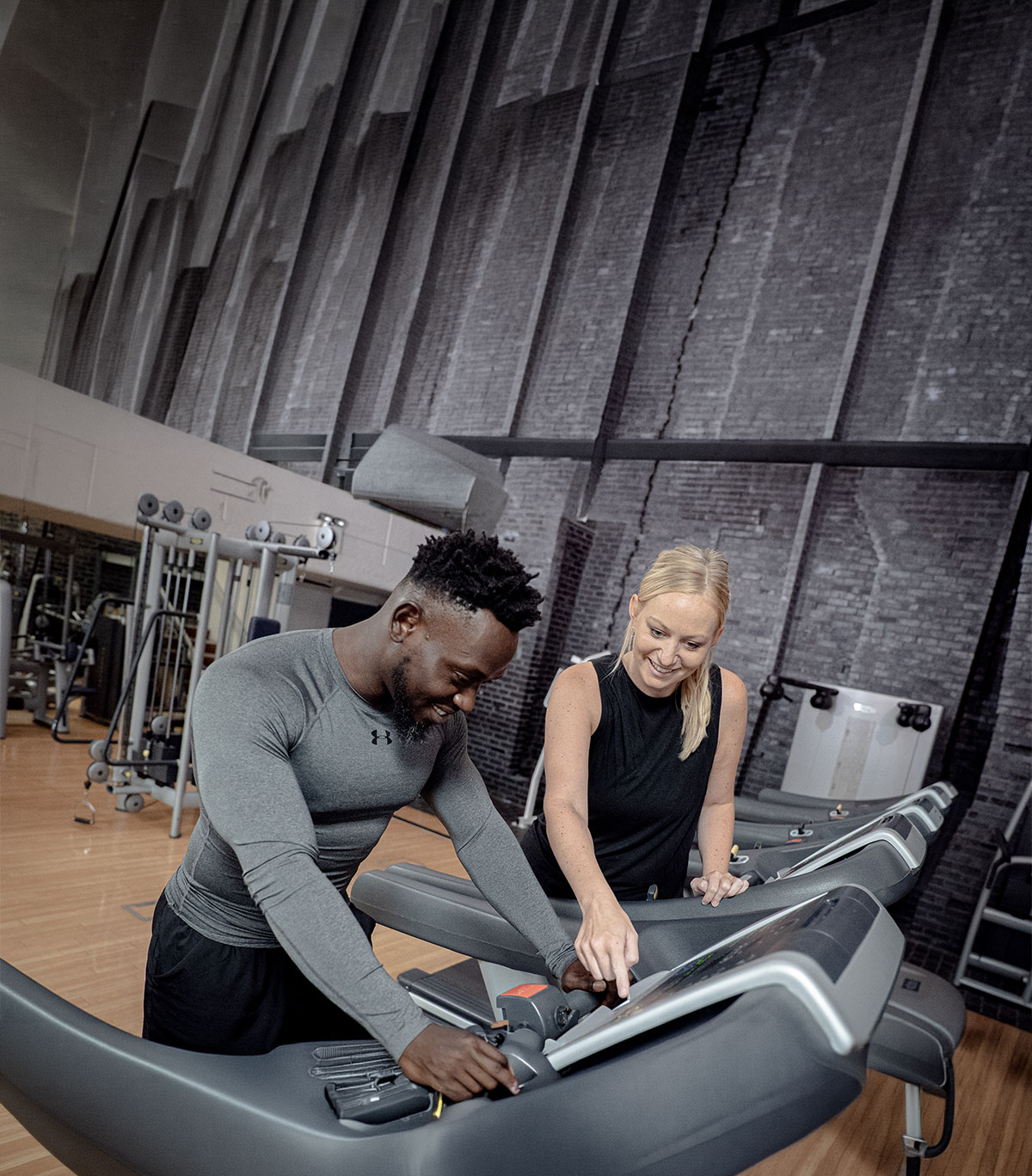 Two people using and adjusting treadmill controls in a gym with exercise equipment and brick walls in the background.