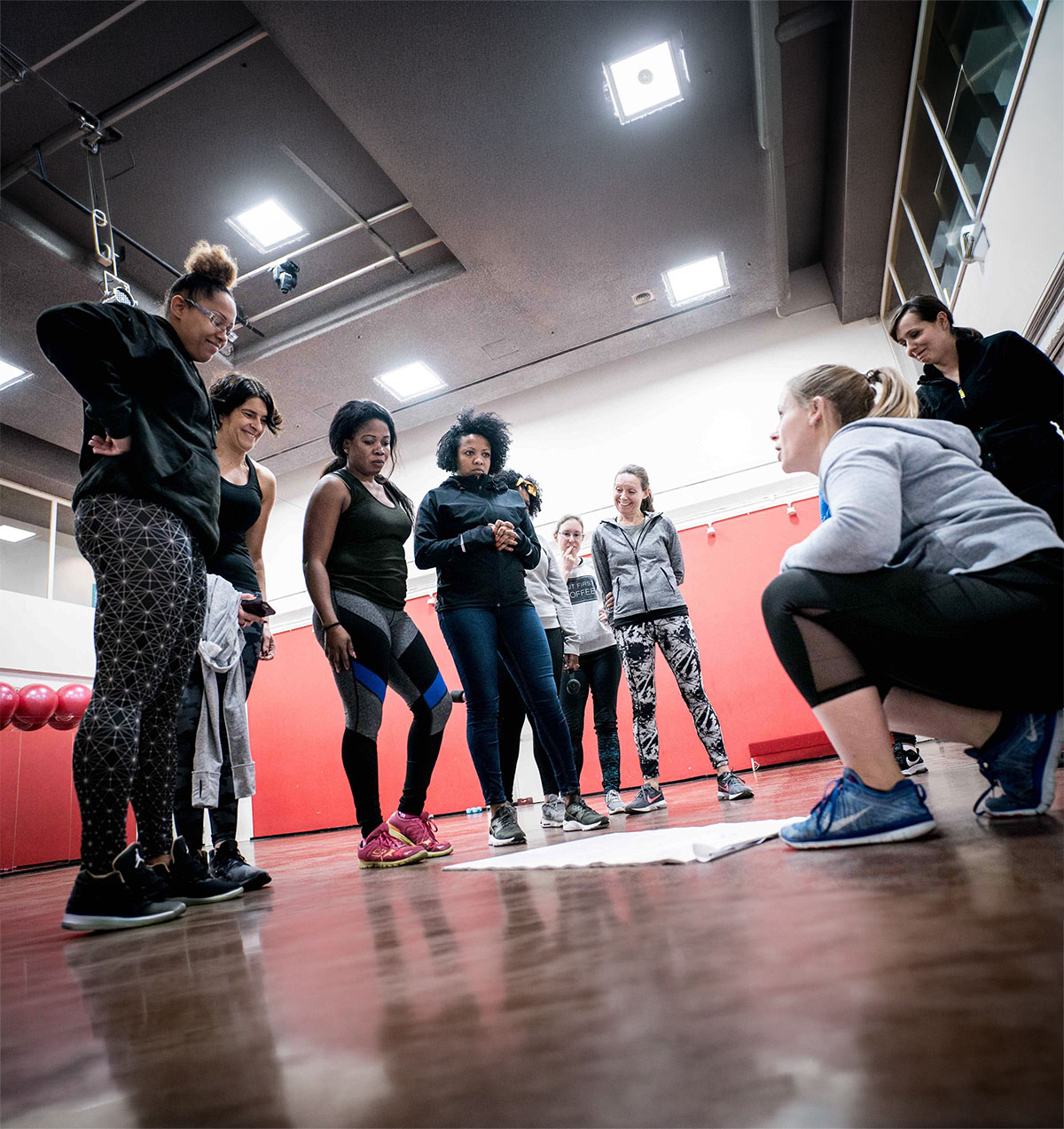 Group of women in workout attire gathered inside a gym, attentively discussing a plan on a paper on the floor.