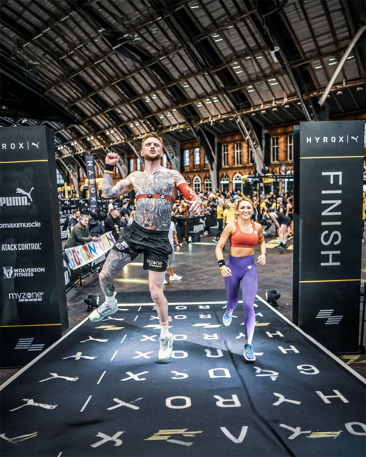 A man and woman cross a race finish line indoors. The man jumps excitedly with a triumphant expression, while the woman smiles, both dressed in workout gear.