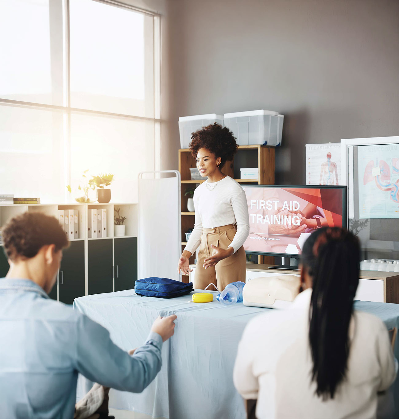 Instructor demonstrating first aid training techniques to students using a CPR mannequin and medical equipment in a classroom.
