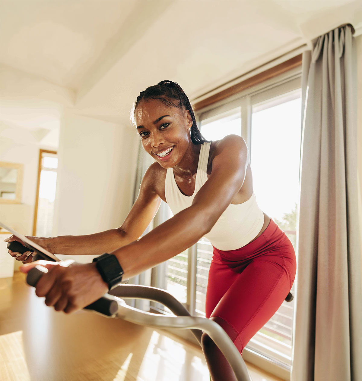 Woman in red leggings and white tank top exercising on a stationary bike indoors near a large window with curtains.