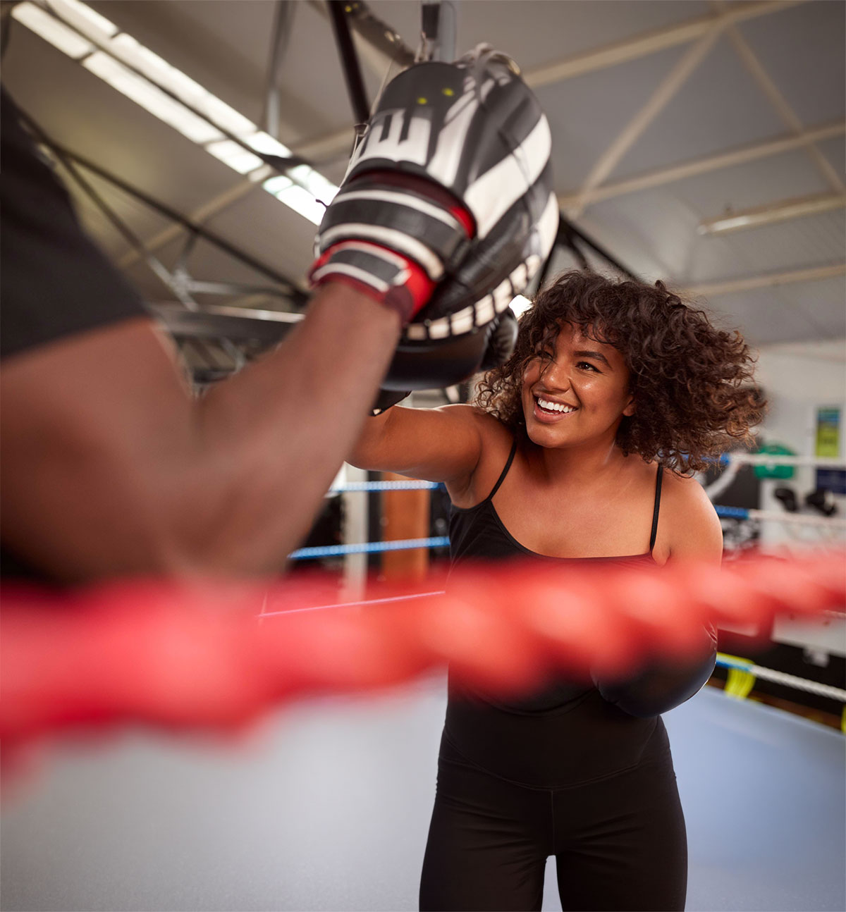 Smiling woman in a boxing ring trains with a partner using focus pads. The atmosphere is energetic and empowering, with bright lighting.