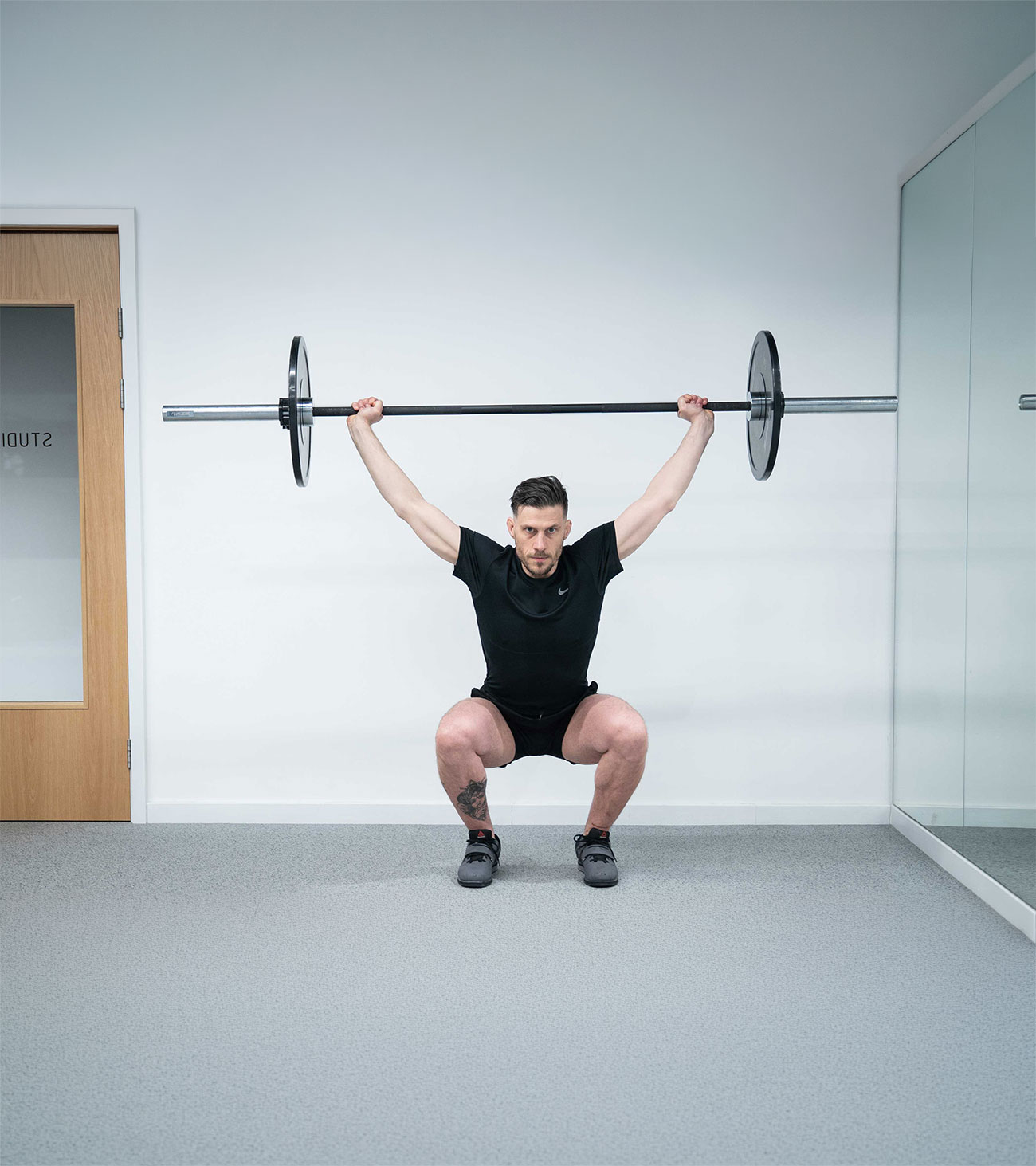 Man in black sportswear performing overhead squat with barbell in a minimalistic gym setting.