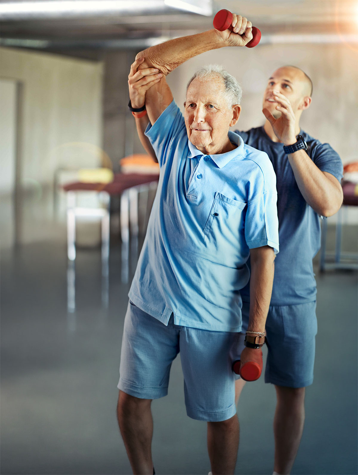 Older man lifting a red dumbbell overhead while being assisted with arm stretch by a trainer in a gym setting.