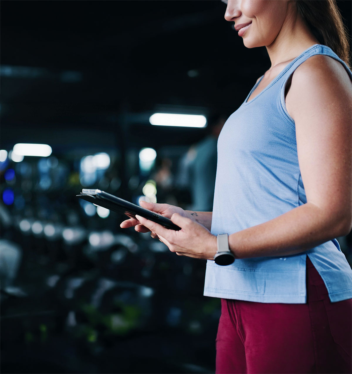A person in a blue tank top stands in a gym holding a tablet, wearing a smartwatch. The blurred background shows gym equipment, creating a focused, active atmosphere.